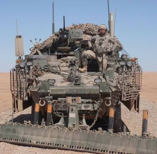 Jonathan is wearing full military camo uniform. He stands with his hands on his hips and a foot up on the back of a military vehicle.