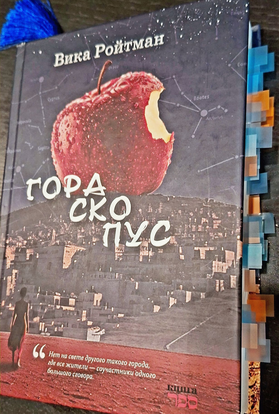 Фото книги Вики Ройтман «ГОРАСКОПУС» с множеством закладок. На обложке изображено надкусанное яблоко на фоне ночного Иерусалима и созвездий.