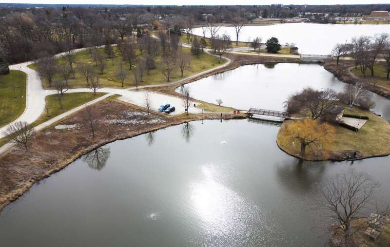 Aerial view of a park with multiple lakes, curving paths, leafless trees, a bridge, and a small island. Overcast sky reflects on the water.