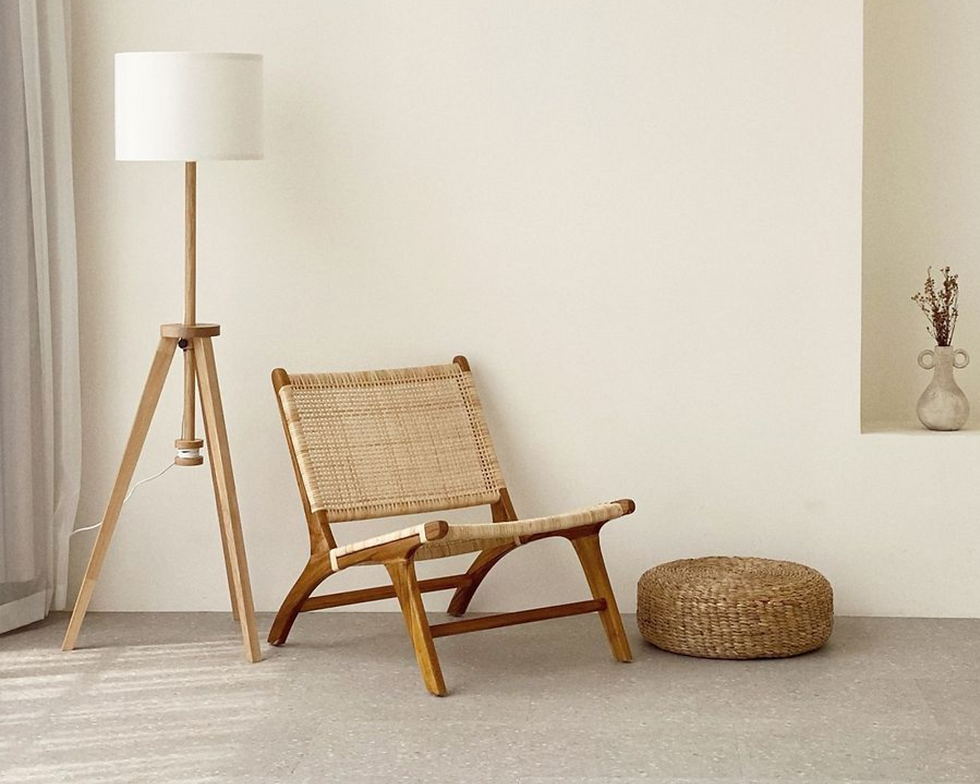 Minimalist room with wicker chair, floor lamp, and round woven pouf. Beige walls and a vase with flowers on a shelf create a calm vibe.