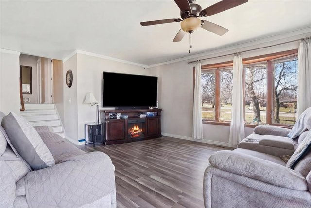 Bright living room with wood-look flooring, large front window, ceiling fan, and stair access.