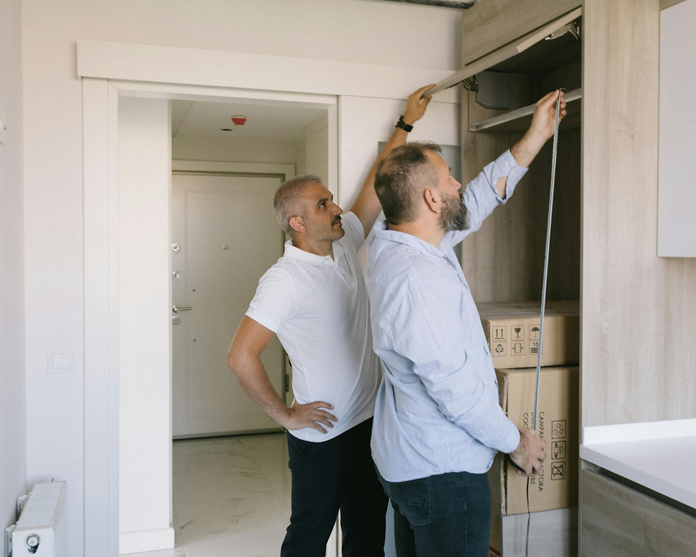 Two men measuring a kitchen cabinet. One holds a tape measure, the other observes. Cardboard boxes in the background, neutral-toned kitchen.