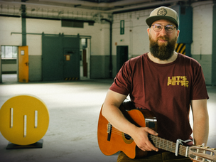 Musiker Wolf Hildebrandt mit Brille, rotem Shirt, langem Bart, Gitarre in der Hand in einem ehemaligen Flugzeughangar auf dem Fliegerhorst Gelände in Oldenburg bei der Aufzeichnung der Kulturschnack Acoustic Sessions