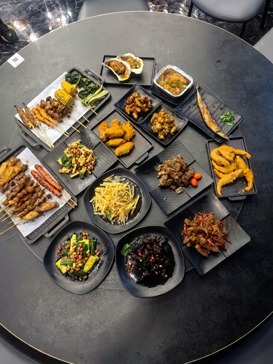 Overhead shot of a round restaurant table filled with many colorful Chinese dishes and skewers on black plates, vibrant food colors against a dark table, portrait orientation.