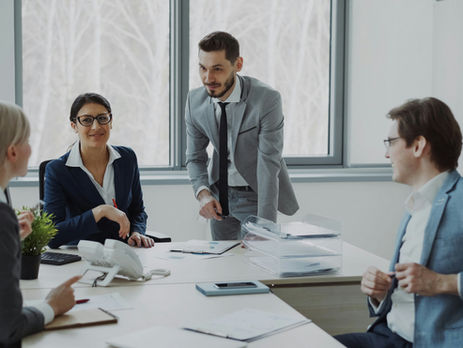 Business meeting with five professionals in suits, smiling and discussing in a bright office with large windows. Papers and devices on table.