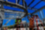 View of two people in ladders, next to curved framework. Deep blue, clear sky in the background.