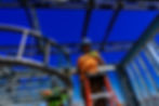 View of two people in ladders, next to curved framework. Man in orange shirt is making a positive hand gesture. Deep blue, clear sky in the background.