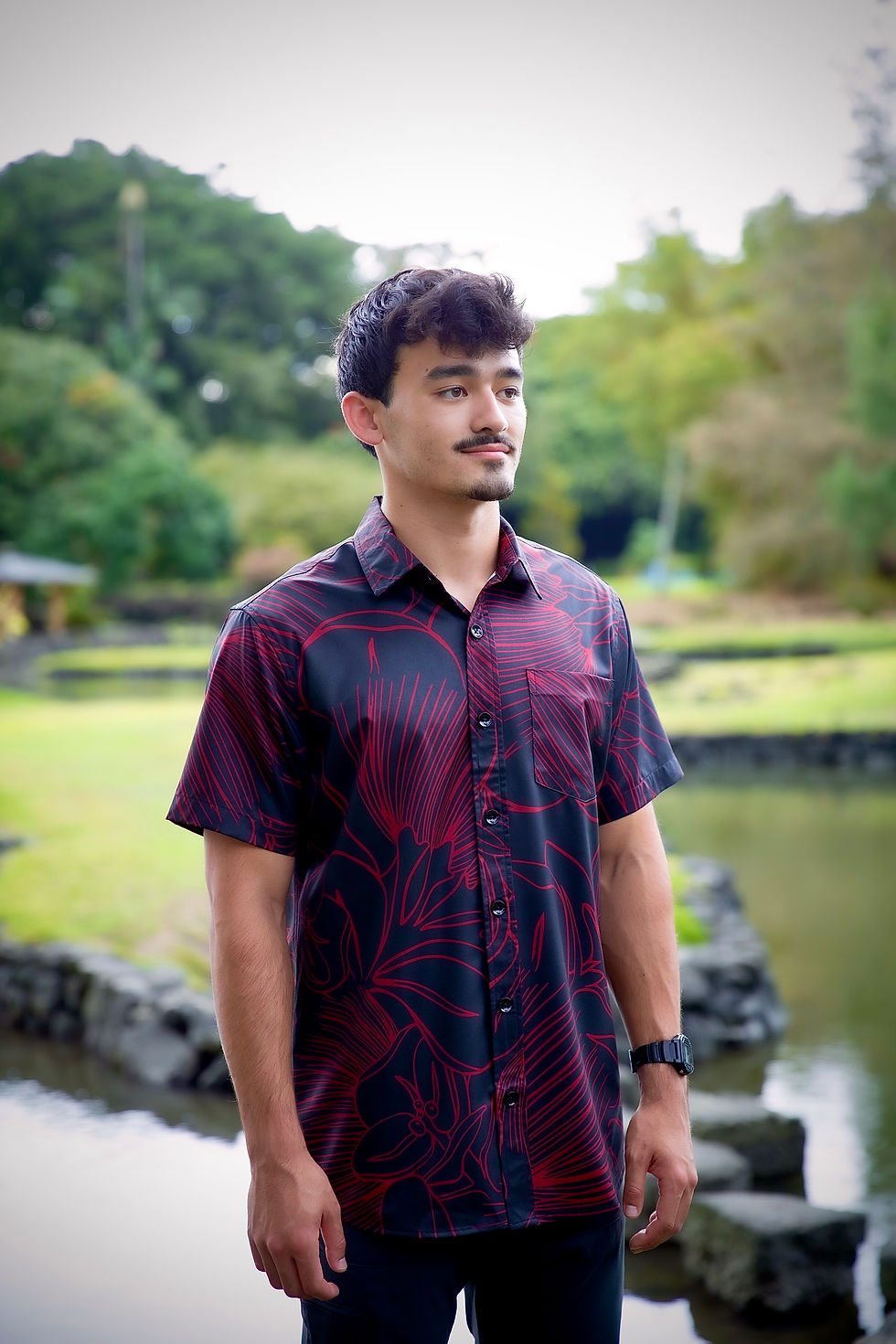 A handsome young man standing in Liliuokalani Park in Hilo, Hawaii wearing a red and black Aloha Shirt with a Lehua Blossom design.  