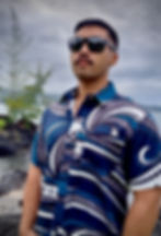A photo of a young man wearing an Aloha Shirt.  The shirt design is a mix of blue, white and gray waves, with imbedded tribal patterns in a repeat pattern design. The photo is taken at Liliuokalani Gardens in Hilo, Hawaii. 