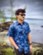A handsome young man standing on a lava rock, standing in front of Hilo Bay.  He is wear a blue Koa leaf and branches hand-drawn repeat pattern design.  