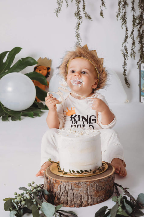 jongen met krulletjes tijdens cake smash met witte verjaardagstaart op boomschijf – curly-haired toddler in cake smash session with rustic white cake