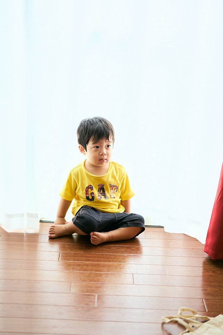 白いカーテン前の木の床に座る幼い男の子。黄色いGAPのTシャツと黒いズボンで、静かに外を見ている。