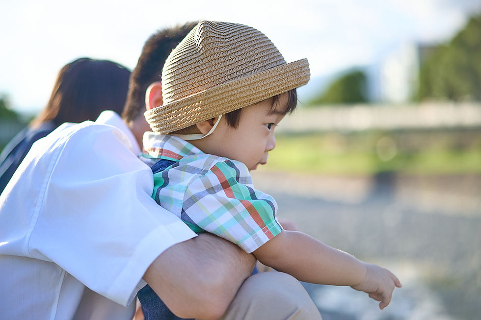 麦わら帽子の子どもを抱く大人の横顔。チェック柄シャツの幼児が前を指さし、屋外の明るい公園で穏やかな雰囲気。