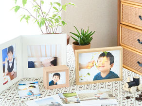 木目の棚と花柄クロスの上に、笑顔の子どもの写真を入れた額やアルバムが並び、観葉植物と木製おもちゃが置かれた明るい室内
