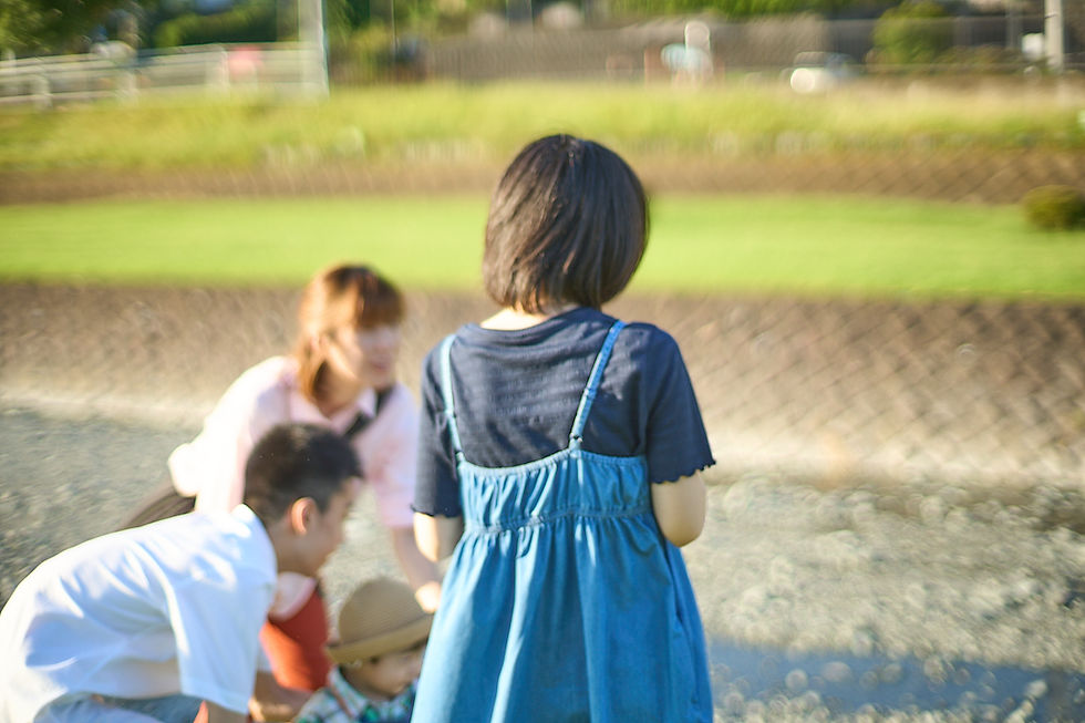 川辺の河川敷で、青いワンピースの少女を中心に大人と子どもたちがしゃがんで集まっている、柔らかくぼけた屋外の光景
