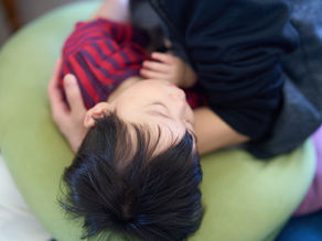大人の膝にもたれて眠る幼い子ども。緑のクッションに頭を預け、赤い縞模様の服を着て安心した雰囲気