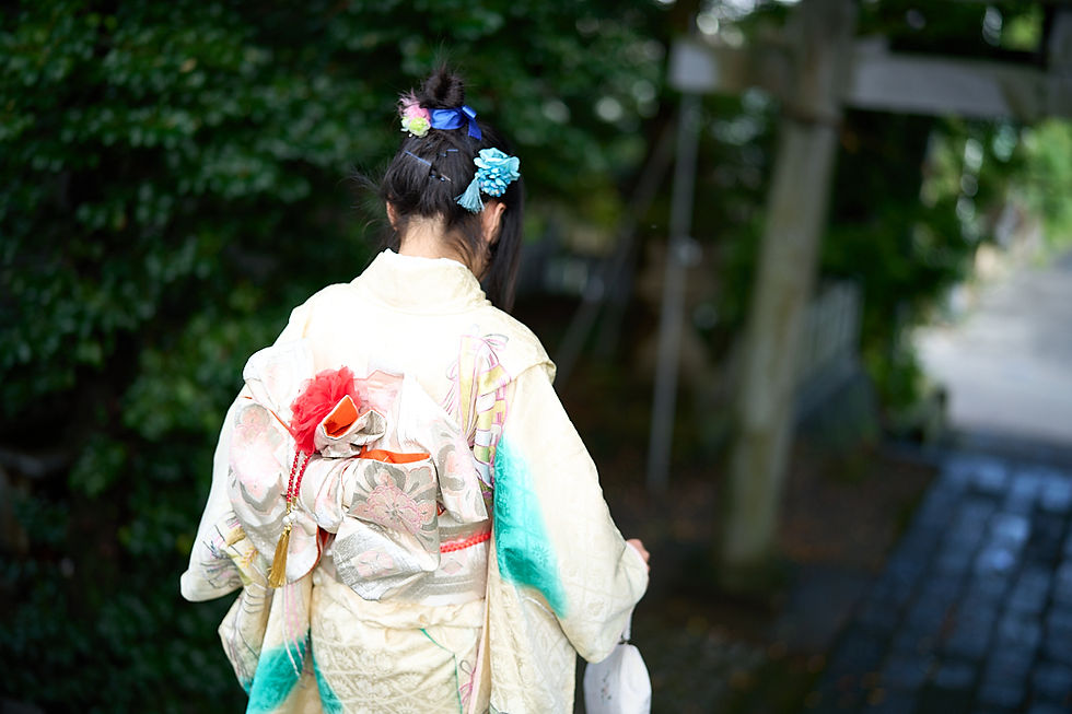 神社の坂道で後ろ姿の少女が、白地に花柄の着物と大きな帯を着け、髪飾りを付けて歩いている。