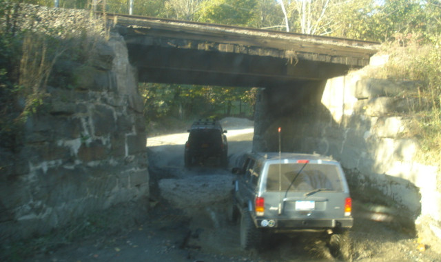 Two Jeeps traversing muddy path under bridge