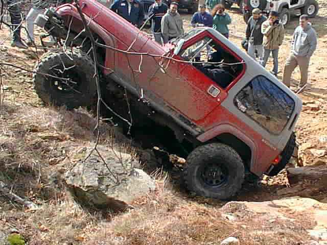 Red Jeep struggling on rocky terrain; onlookers watch