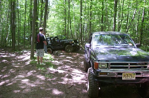 Two men watch as a truck tows a Jeep stuck in the woods