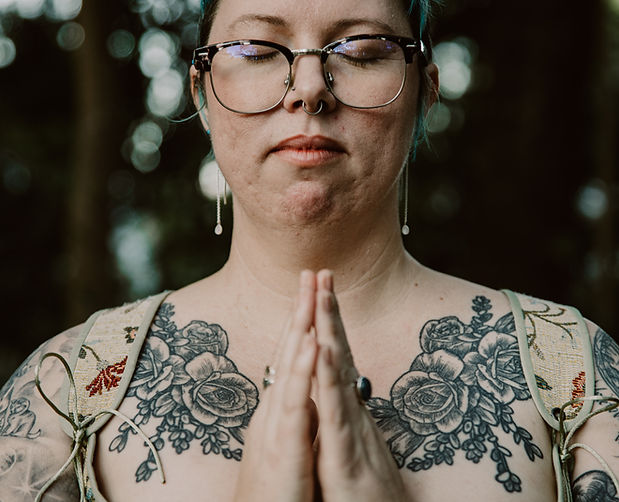 Brogey, a caucasian person with glasses and tattoos faces the camera, eyes closed, hands in anjala mudra.