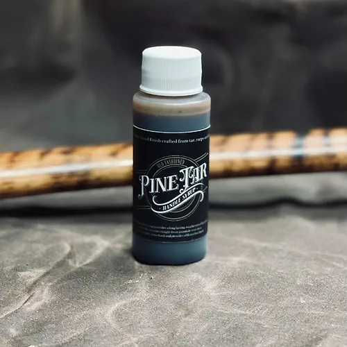 A small bottle of pine tar handle syrup sits on a wooden surface. The syrup is a dark brown color and has a thick, syrupy consistency. A refinished axe handle is laying in the background. The axe handle is made of wood and has a dark brown finish. The syrup has coated the axe handle, giving it a protective and weather-resistant finish.