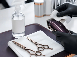 Gloved hands disinfecting clipper blades on a salon station beside shears, a folded white towel and an unbranded spray bottle.