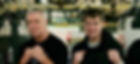 Two men boxing in a gym, one with grey hair and a black shirt, the other with brown hair and a black hoodie