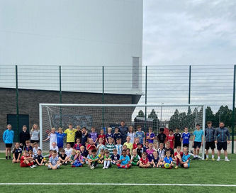 Group of young soccer players, coaches, and volunteers pose for a photo on a green field in front of a soccer goal.