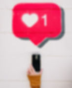 Hand holds a smartphone against a white brick wall with red heart and "1" notification graffiti, suggesting a social media like.
