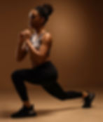 Woman in workout attire performs a lunge in a warm, brown-lit setting. She appears focused, wearing a sports bra and black leggings.