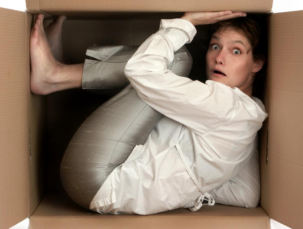 Person in white shirt crouched in a cardboard box, looking surprised. Box is large but cramped, suggesting a humorous or uncomfortable situation.