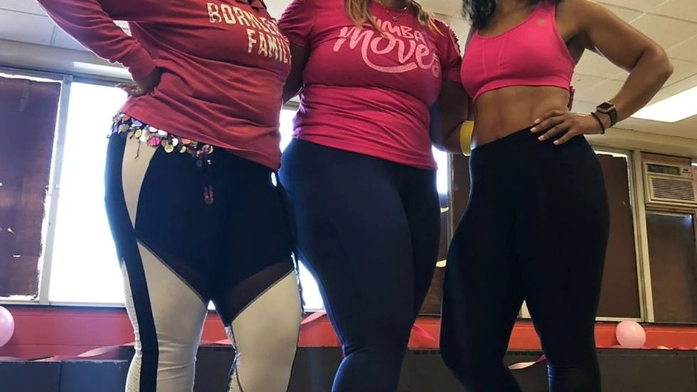 Three women in a dance studio wearing pink tops and black leggings laugh together. Shirts read "Zumba moves" and "Bombafam." Bright, cheerful mood.