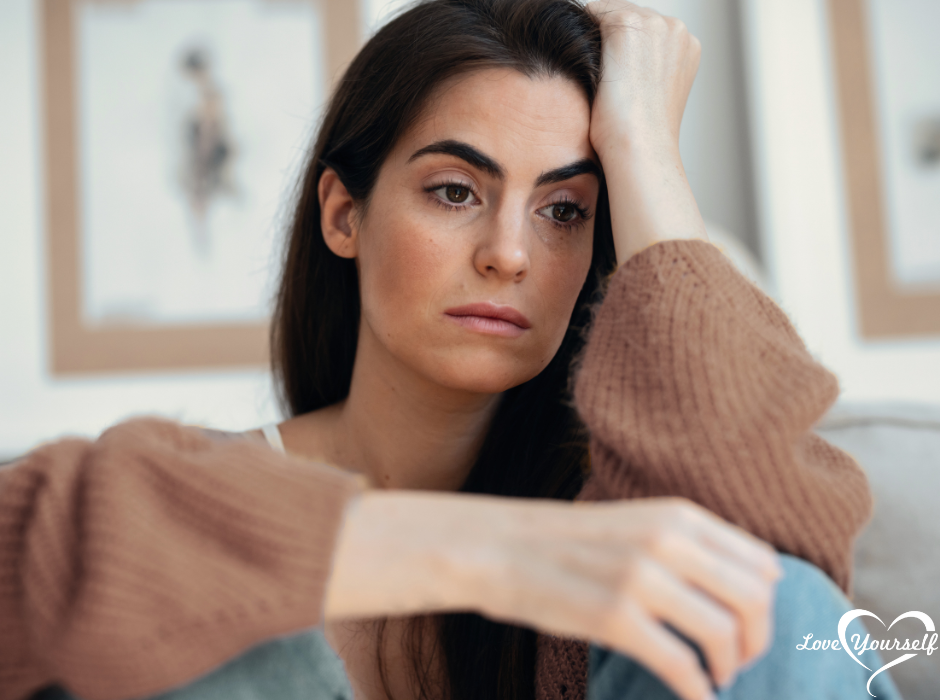 Woman with long hair looks pensive, resting head on hand, wearing a brown sweater. Background has blurred wall art. Text: "Love Yourself".