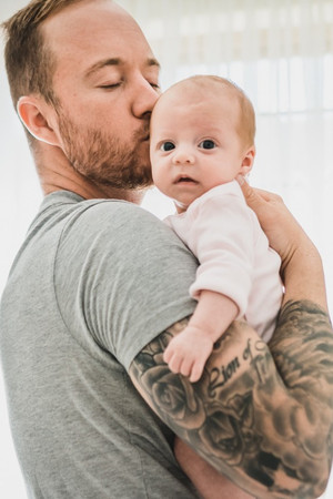 father holding newborn baby and kissing her head. baby's hand hangs over tattooed arm of her dad. newborn lifestyle photography melbourne