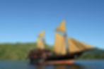 A magnificent traditional wooden sailing ship with unfurled beige sails, anchored in calm turquoise waters with lush green hills in the background under a clear blue sky.