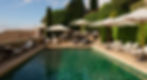 A serene outdoor swimming pool surrounded by elegant lounge chairs and umbrellas, set against a backdrop of lush green terraced gardens and cypress trees under a bright sky.