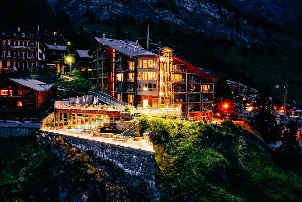 A beautifully illuminated luxury hotel perched on a hillside in an alpine village at night, with warm lights glowing from its windows against a backdrop of dark mountains.