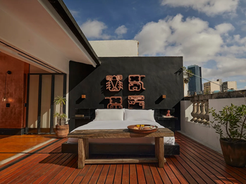 Open-air rooftop bedroom at La Valise Mexico City featuring a white bed on a wooden deck terrace, decorative copper art mounted on a black wall behind the headboard, potted plants, and the city skyline under a partly cloudy sky.