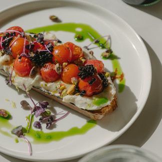 A vibrant bruschetta-style toast topped with cherry tomatoes, cheese, herbs, and a drizzle of green oil, served on a white plate.