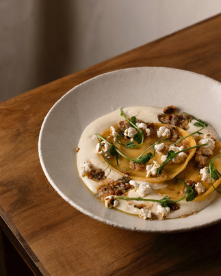 A gourmet dish of pasta, possibly ravioli or tortellini, topped with a creamy sauce, herbs, and ground ingredients, presented on a white plate.