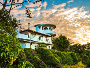 A unique, multi-level white building with rounded architectural elements, nestled among lush green foliage under a dramatic sunset sky.