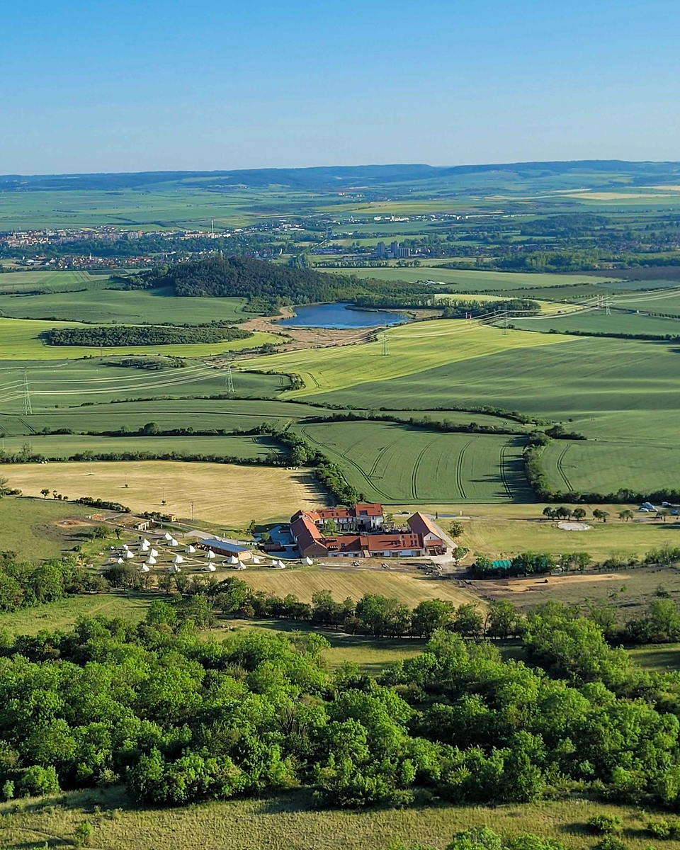 Aerial view of Oblík Resort surrounded by regenerated habitat, featuring expansive native grass meadows, wildflower fields, a small natural pond, and the restored farmhouse set against rolling green hills.