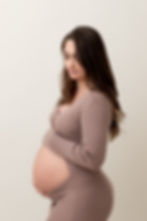 Expecting mother with long brown hair standing on a cream backdrop in a mocha colored dress looking down toward her belly.