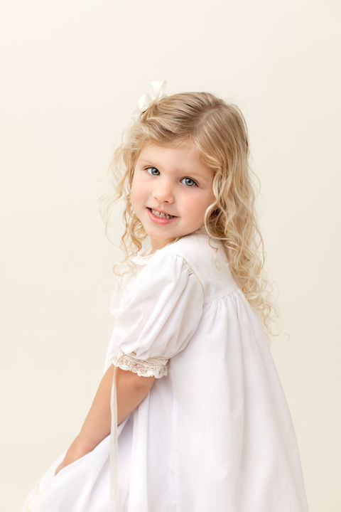 Child portrait of a girl with long blonde hair in a white heirloom dress sitting and smiling. 