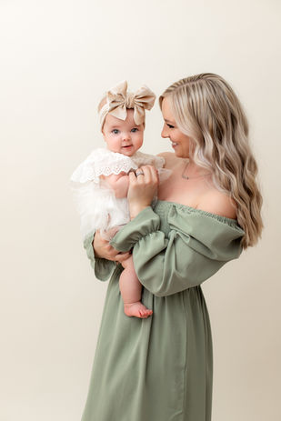 Motherhood studio session portrait of a mother with long blonde hair in a green dress holding her baby and looking at her while baby looks at the camera.