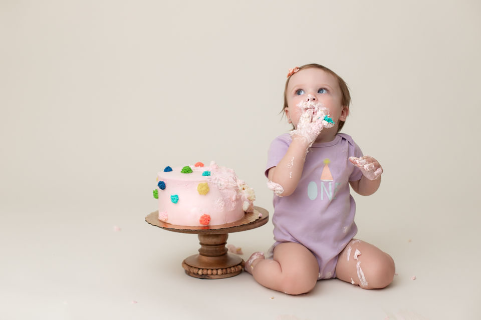 Child portrait of a one year old girl in a purple onesie eating her cake at her cake smash session.