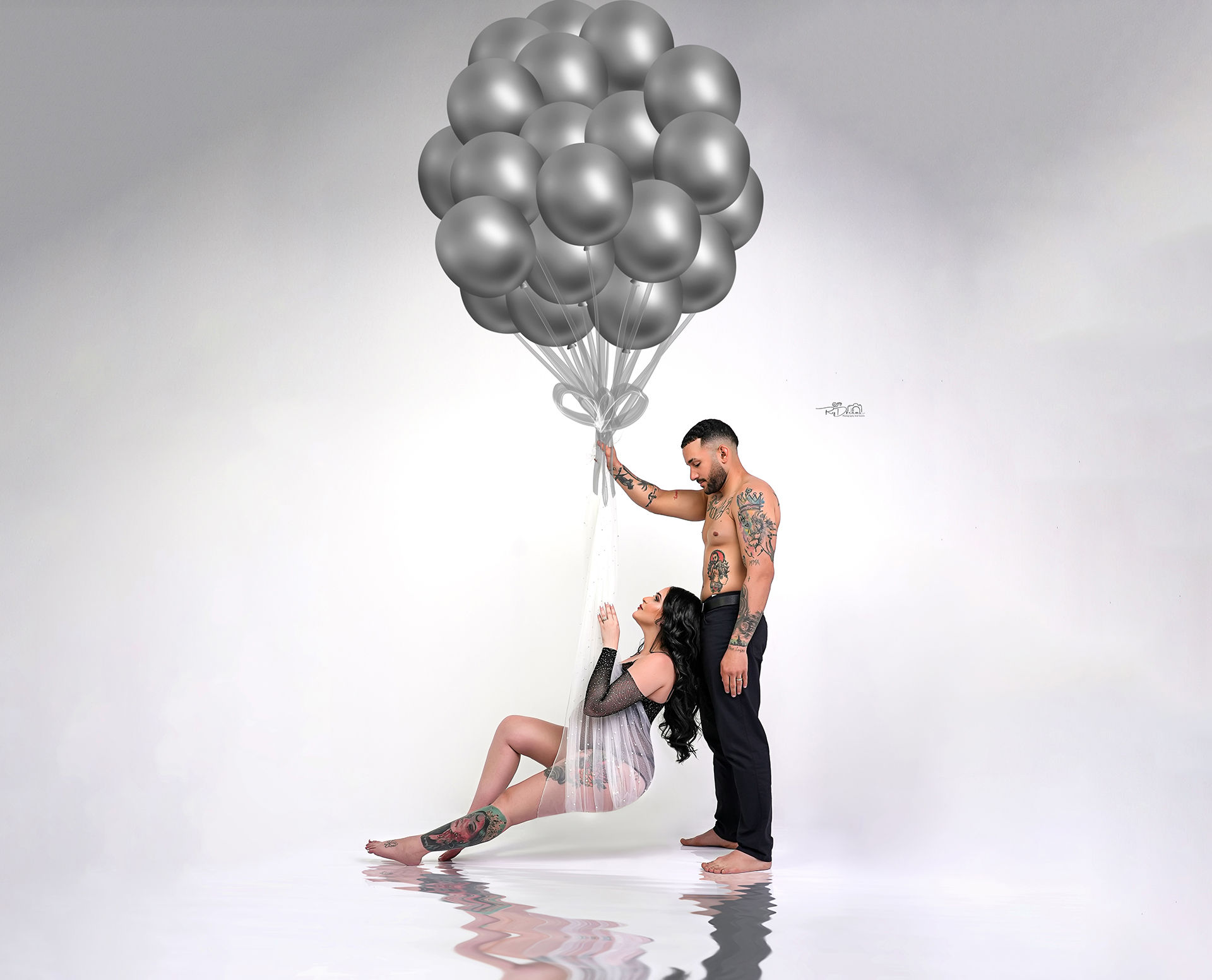 Pregnant couple in an in-studio session, with the husband holding his wife lovingly while she holds a colorful bouquet of balloons, celebrating their upcoming arrival.