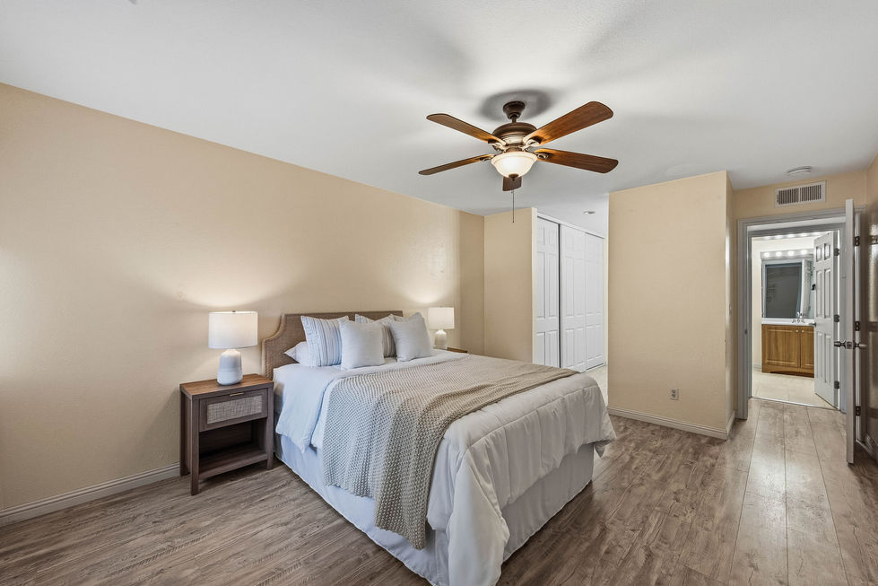 Modern bedroom with white bedding, wood‑tone flooring, nightstands, ceiling fan, and hallway leading to bathroom and closet.