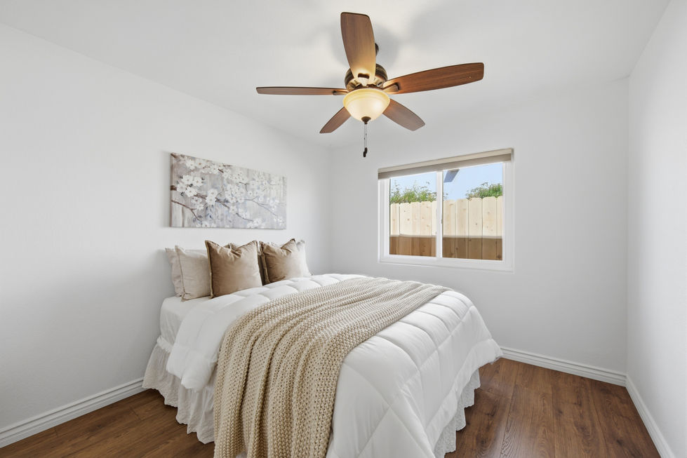 Guest bedroom with white bedding, beige accents, wood flooring, ceiling fan, and window with backyard view.